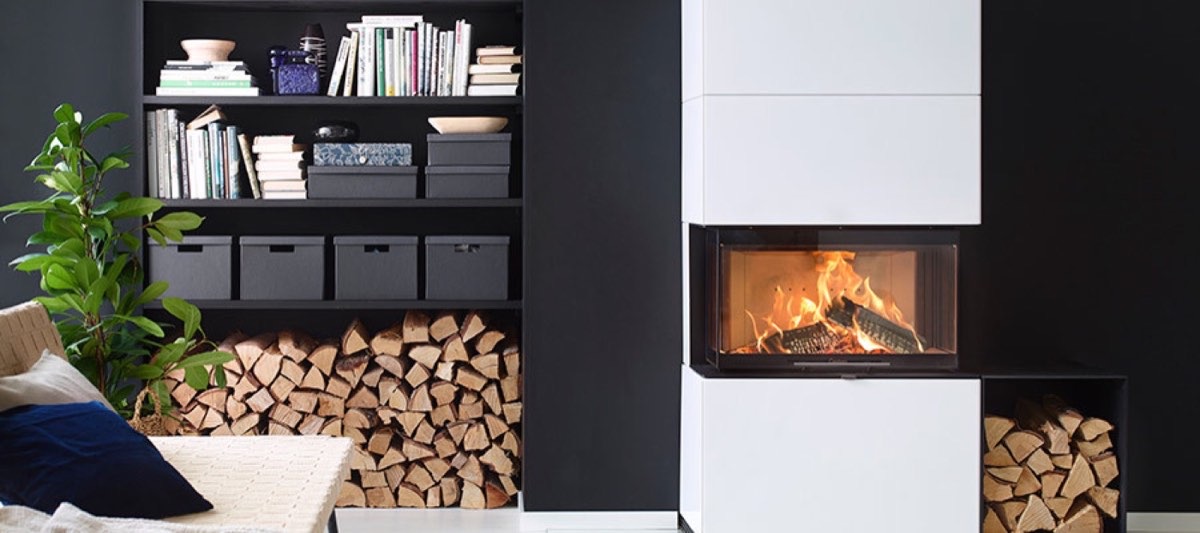 Weißer Heizkamin mit breiter Feuerscheibe und brennendem Holz, eingebaut in eine Wohnwand mit Bücherregal und Holzlager